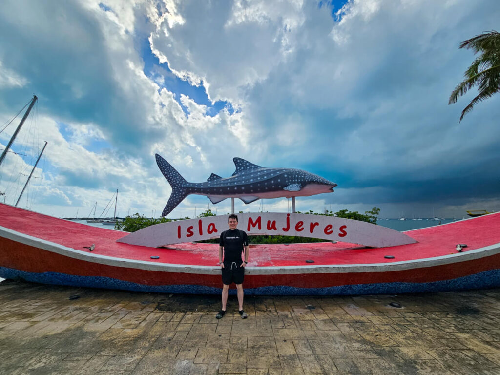 Dan stood in front of the famous whaleshark Isla Mujeres sign. The sky is moody and the ground is wet from rain.