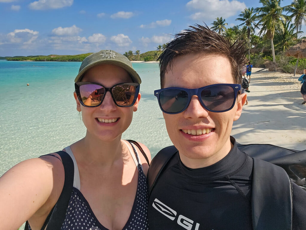 Dan and Lucy standing smiling on a white sand beach on Isla Contoy. Palm trees are moving in the wind and the coast is arching into the distance.